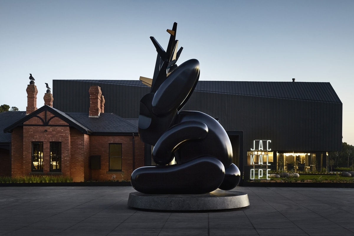 A large dark sculpture in front of a small brick house and large modern structure,
